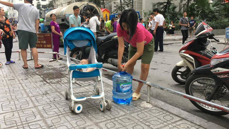 Người mẹ trẻ một tay đẩy xe cho con, một tay xách can nước 20 lít ở chung cư Linh Đàm (Ảnh GT)